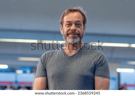 Street portrait of a happy 50-55 years old man with beard in gray t-shirt on blurred background of train station. Concept: tourist or traveler, shopper or actor, loader or military pensioner. Royalty-Free Stock Photo #2368559245