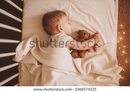 A little baby in a white bodysuit sleeping in a white crib holding a teddy bear and surrounded by small lights Royalty-Free Stock Photo #2368574225