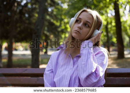 Young blonde European woman sits on a bench in the park and listens to music on headphones, pressing them with her hand to her head and looking sadly to the side