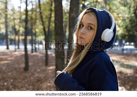 Cute blonde girl in a dark jacket with a hood on and white headphones on her head looks at the camera holding her hair in hand in an autumn park against the background of trees