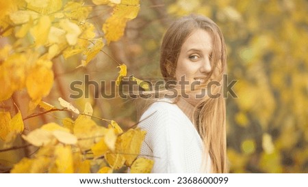Beautiful young woman with light brown hair in a white sweater on a background of foliage in an autumn park. Portrait of an attractive happy woman among colorful yellow leaves. Fall season. Autumn.