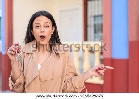 Young woman at outdoors holding home keys at outdoors surprised and pointing side