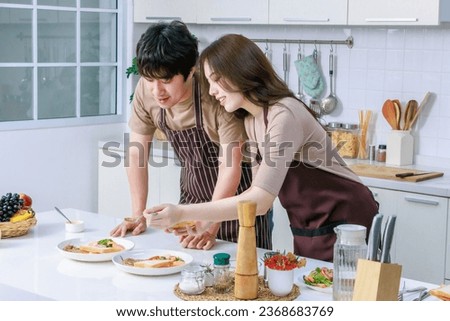Asian young lover couple husband and housewife in casual outfit with striped apron standing using spoon testing cooking breakfast meal in full decorated modern kitchen with ingredients equipment.