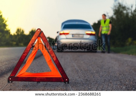 Unrecognizable sad driver in reflective vest. Male driver standing near broken car with open up hood. Red triangle to warn other road users of car breakdown or engine failure stop at countryside