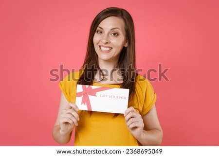 Smiling beautiful attractive young brunette woman 20s in yellow casual t-shirt posing standing holding in hands gift certificate looking camera isolated on pink color wall background studio portrait