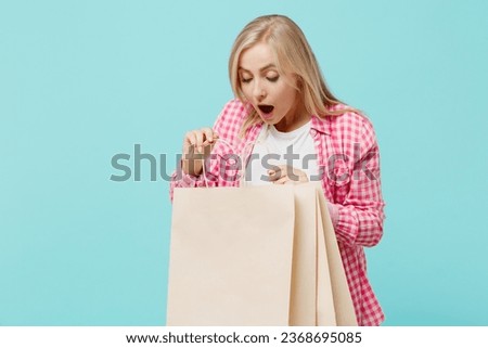 Young amazed woman she 30s in pink shirt white t-shirt hold look at package bags with purchases after shopping isolated on plain pastel light blue background studio portrait. People lifestyle concept