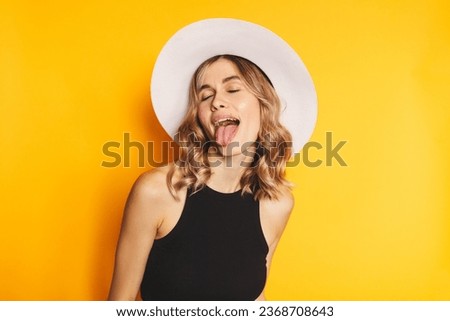 Portrait of naughty positive woman with wavy hair in white hat standing with closed eyes and demonstrating tongue, expressing disobedience disrespect, teasing grimace studio shot, yellow background. Royalty-Free Stock Photo #2368708643
