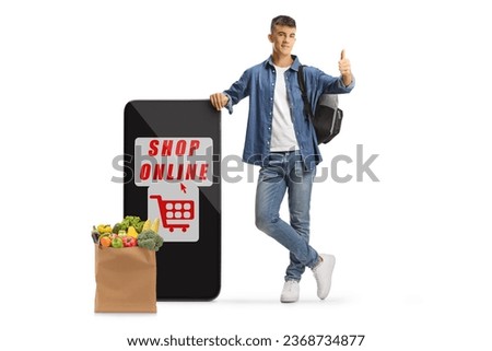 Male student gesturing thumbs up and leaning on a mobile phone with online shopping app and a grocery bag isolated on white background