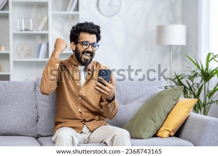Young man received an online notification of a winning message sitting at home in the living room on the sofa, the winner is satisfied with results of the achievement joyfully holding his hand up. Royalty-Free Stock Photo #2368743165