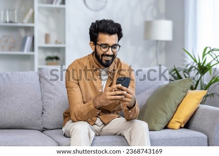 Young hispanic man sitting at home on sofa in living room of house, happy and smiling man holding phone, using smartphone app, typing message and watching video, online shopping. Royalty-Free Stock Photo #2368743169