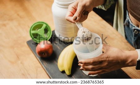 Woman in jeans and shirt with a measuring spoon in her hand puts portion of whey protein powder into a shaker on wooden table with white capsules, bananas and apple. Process of making protein drink. Royalty-Free Stock Photo #2368765365