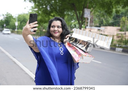 Young indian plus size woman holding shopping bags taking selfie picture with smart phone outdoors on the street. Overweight lady using phone.