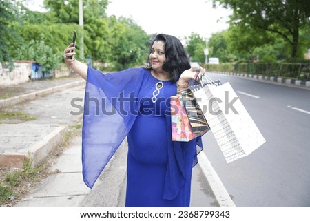 Young indian plus size woman holding shopping bags taking selfie picture with smart phone outdoors on the street. Overweight lady using phone.