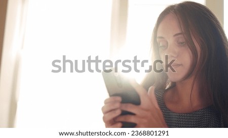 pretty girl looking at smartphone near the window. internet social network concept. girl makes online purchases on a smartphone near the window at home. girl reading news sun on a smartphone
