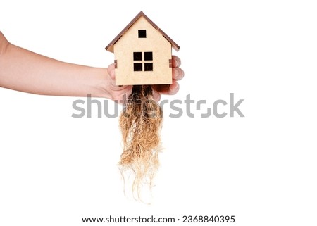 Close-up of a hand holding a small, intricately crafted wooden model house featuring lush, intertwined roots, set against a clean white backdrop. Relocation concept.