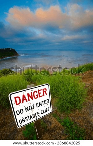 A bold sign, posted above a rocky coastline, warns visitors about the danger of an unstable cliff. A dramatic cloudy sky can be seen in the background. High quality photo