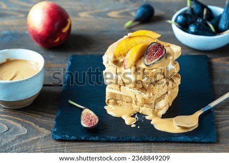 Peanut Butter spread on the toasts, cut figs and peaches on the top, bowl with butter, fruits over wooden background