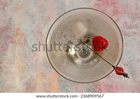 Looking down shot of martini glass with beverage, one ice cube, one raspberry and food pic inside. Placed on pinkish pastel background.  