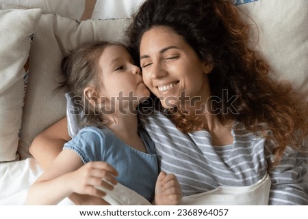 Cute little preschooler daughter and happy millennial mom waking up in cozy home bed together, smiling young mother or nanny relaxing in comfortable bedroom with small girl child cuddling and hugging