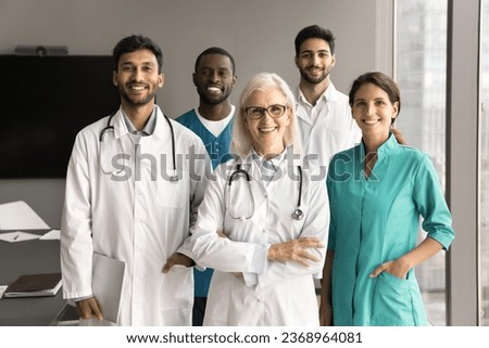 Positive confident senior clinic chief doctor and younger diverse team of practitioners, surgeons, nurses standing together in hospital office, looking at camera with toothy smiles