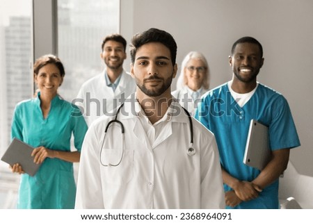 Positive attractive young Arab doctor wearing uniform and stethoscope, looking at camera, standing in front of multiethnic colleagues in hospital office. Medical boss and clinic staff portrait