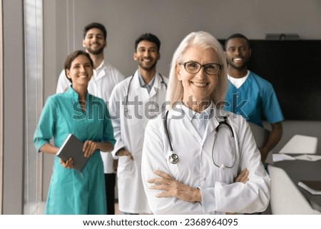 Happy elder mature head doctor woman posing with arms crossed with toothy smile and multiethnic team of younger colleagues, hospital staff in blurred background
