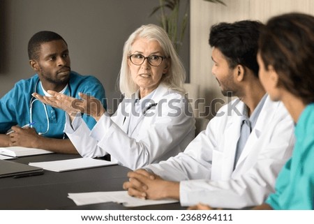 Serious senior head doctor talking to diverse hospital staff at meeting table, speaking, giving instructions, explaining medical problem. Medical expert, mentor training younger interns Royalty-Free Stock Photo #2368964111