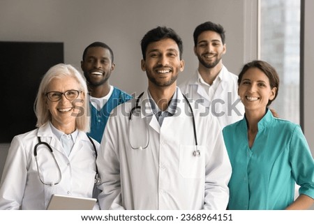 Happy young Indian medical boss and diverse team of doctors, practitioners, surgeons in uniforms looking at camera with toothy smiles, posing for portrait. Multiethnic small clinic staff shot