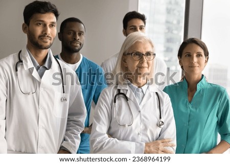 Multiethnic different aged group of doctors standing together for shooting, looking away. Confident senior older head of clinic posing in front of team with hands folded