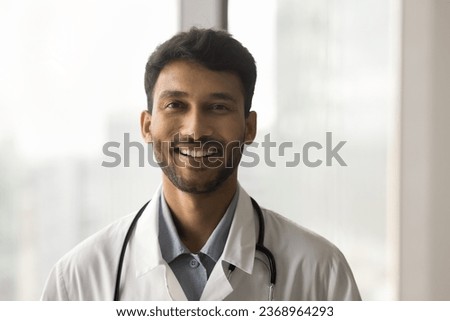 Positive attractive Indian doctor man in white uniform coat and stethoscope head shot portrait. Handsome young male practitioner looking at camera with toothy smile. Video call screen view Royalty-Free Stock Photo #2368964293
