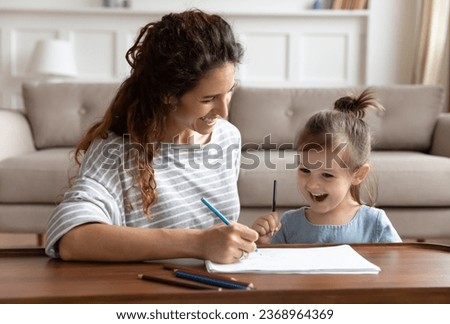 Overjoyed millennial mother and small preschooler daughter relax have fun together in living room drawing pictures, excited young mom or nanny painting with colorful pencils with smiling little girl