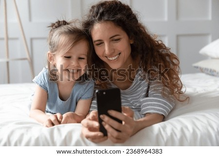 Happy millennial mom and cute little preschooler daughter lying in cozy bed make self-portrait picture on smartphone, smiling young mother and small girl child have fun use cellphone together