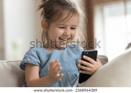 Happy cute little girl sit on couch laugh watching funny video on cellphone, smiling small preschooler kid relax on sofa have fun using modern smartphone gadget, children and technology concept