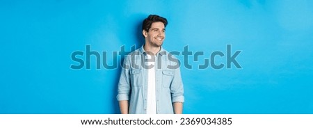 Handsome smiling adult man in casual outfit, smiling and looking left at promo offer, standing against blue background. Royalty-Free Stock Photo #2369034385