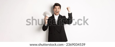 Portrait of handsome businessman in black suit, winning money and rejoicing, raising hand up with excitement, standing against white background.
