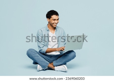 Full body surprised young IT man of African American ethnicity he wear shirt casual clothes sit hold use work on laptop pc computer isolated on plain pastel light blue cyan background studio portrait