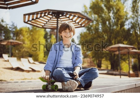 Carefree cute with skateboard. Full length of cheerful teen boy looking at camera with smile while sitting on skateboard against in summer city park