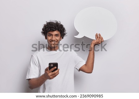 Waist up shot of cheerful handsome Hindu man with glad expression dressed in casual tshirt uses mobile phone for chatting online holds blank speech bubble for your text isolated on white wall