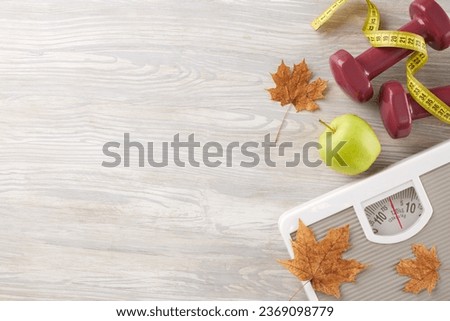 Autumnal body enhancement idea. Top view photo of dumbbells, floor scales, tape measure, fresh apple, dry maple leaves on light wooden background with advertising zone