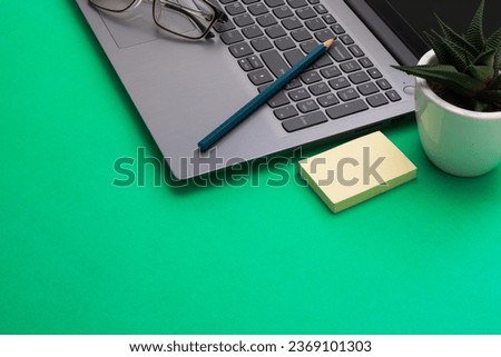 A Silver Gray Laptop with Eyeglasses, Pencil, Sticky Notes, and a Planter on a green Background. Front view.