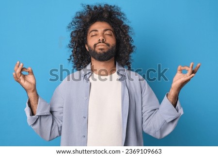 Young relaxed bearded Arabian man takes yoga pose and meditates to clear head of negative thoughts and plunge into zen trance dressed in casual clothes stands on plain blue background.