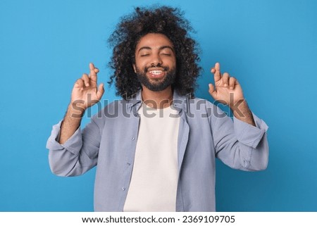 Young cheerful superstitious Arabian man clasps fingers in cross after making wish and closes eyes with smile in anticipation of birthday surprise or dream come true stands posing in blue studio.
