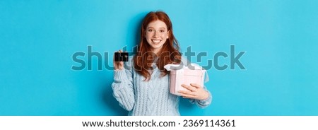 Happy redhead girl buying gifts with credit card, holding box with present and smiling, standing over blue background.