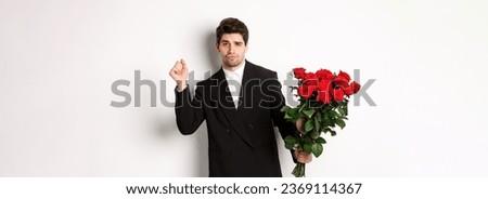 Image of elegant and sassy man in black suit, looking confident and holding bouquet of red roses, going on a romantic date, standing against white background.