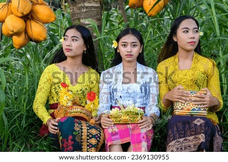 Three Balinese teenagers in traditional outfits resting in the shade of a coconut palm tree.
