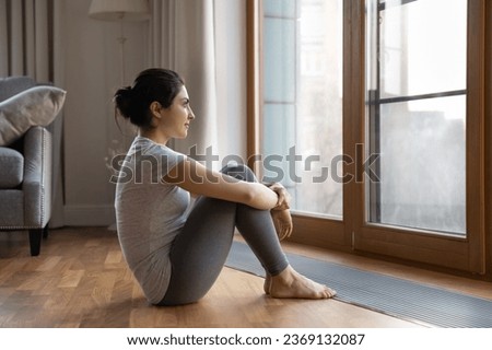 Dreamy Indian woman looking out window, sitting on warm wooden floor at home, pensive young female in grey sportswear dreaming of good future in living room, visualizing success, new opportunities Royalty-Free Stock Photo #2369132087