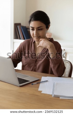 Vertical photo of confident Indian woman using laptop, sitting at office desk, focused young businesswoman professional or freelancer looking at computer screen, working on project online or studying