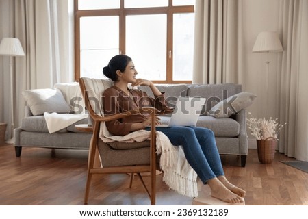 Smiling dreamy Indian woman using laptop, sitting in armchair at home, looking in distance, happy thoughtful young female freelancer looking in distance, dreaming of good future, visualizing success Royalty-Free Stock Photo #2369132189