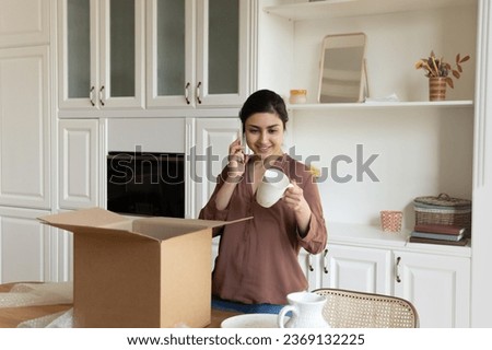 Smiling Indian woman talking on phone, chatting with friend, unpacking box with fragile online store order, holding porcelain cup, happy young female unboxing parcel with belongings in new apartment Royalty-Free Stock Photo #2369132225