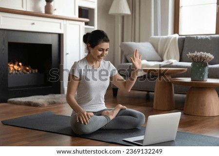 Smiling Indian woman waving hand using laptop webcam starting online yoga lesson, greeting teacher trainer, happy young female in sportswear sitting in lotus pose, working out doing exercises at home